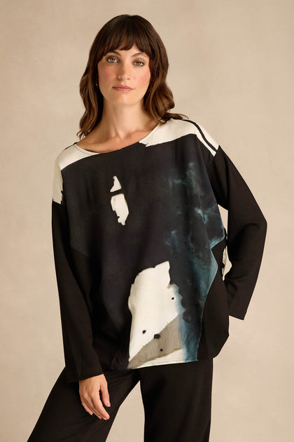 A woman stands against a neutral background, wearing the Abstract Watercolour Scoop Neck Top—a long sleeve, relaxed fit black top featuring an abstract black, white, and gray watercolor pattern—paired with coordinating black pants.