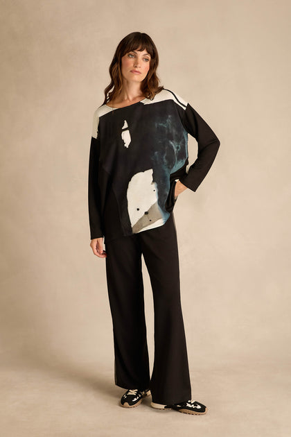 A woman stands facing forward against a neutral studio backdrop, wearing black wide-leg pants, black loafers with white detailing, and the Abstract Watercolour Scoop Neck Top—a long sleeve relaxed fit top.
