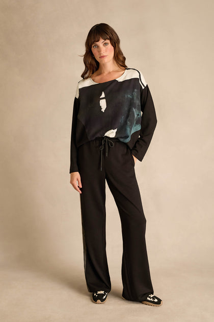 A woman stands against a neutral background wearing a black and white abstract print top, paired with the Side Stripe Wide Leg Trousers in black featuring a comfortable drawstring waist, and black shoes with a white pattern.