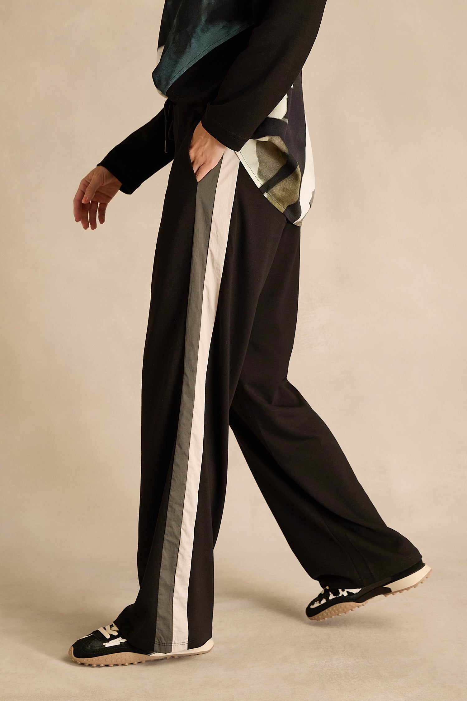 Wearing the Side Stripe Wide Leg Trousers, a black long-sleeve top, and black sneakers, this person stands against a beige background.