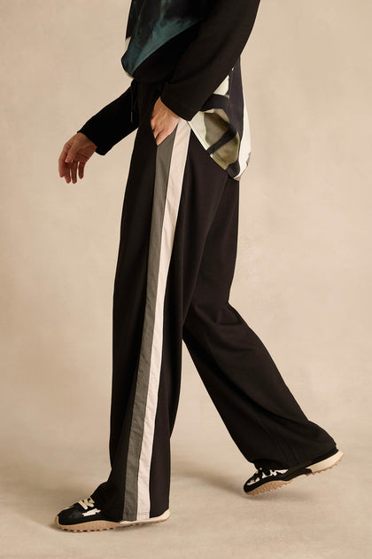 Wearing the Side Stripe Wide Leg Trousers, a black long-sleeve top, and black sneakers, this person stands against a beige background.