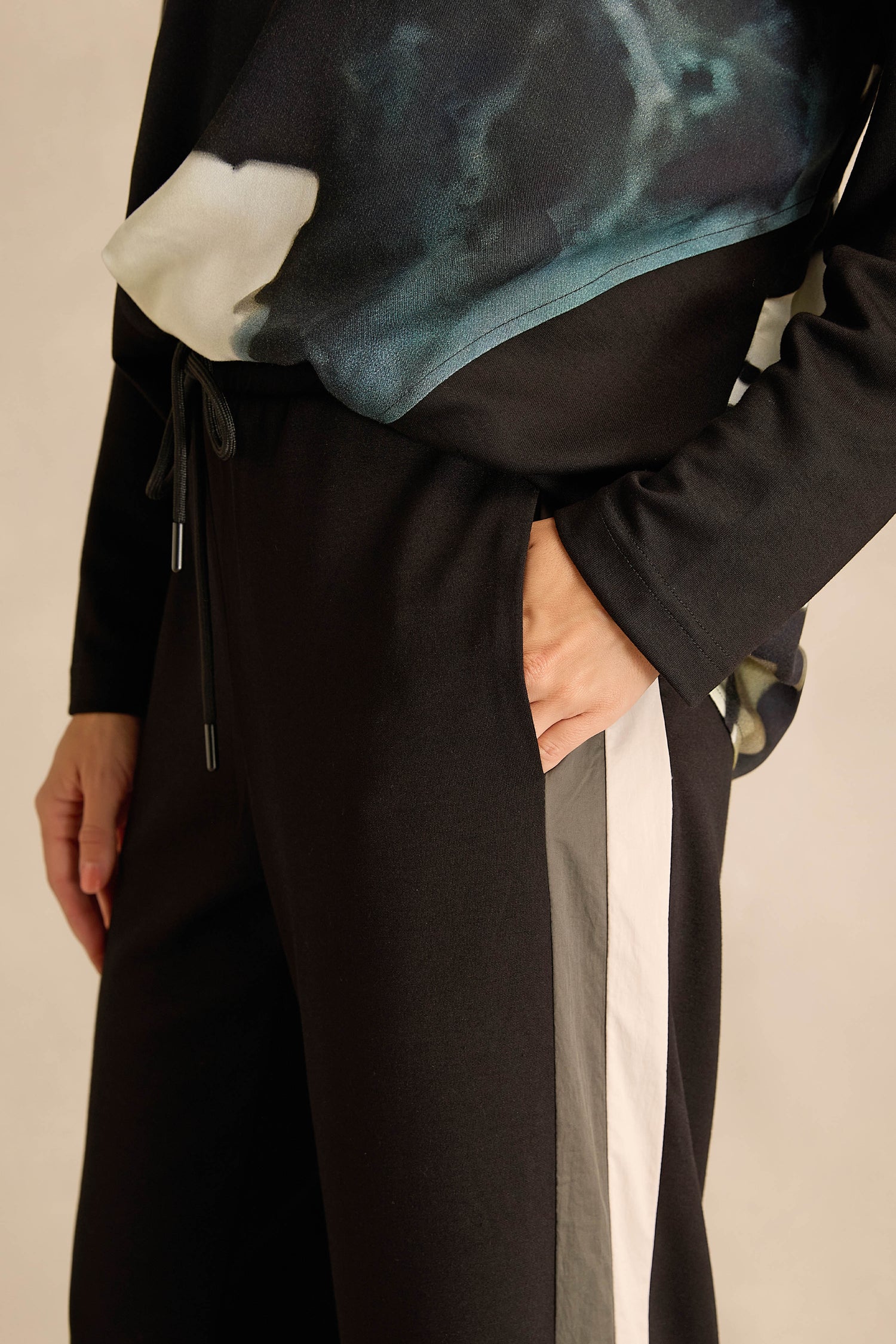 Wearing the Side Stripe Wide Leg Trousers, which feature white and gray side stripes, paired with a patterned black top, this person stands with one hand in their pocket.