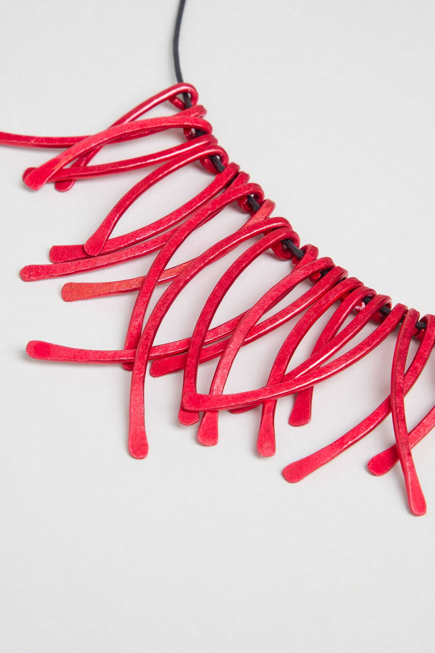 Introducing the Arc Necklace—a contemporary jewellery piece crafted from anodised aluminium, featuring multiple red, curved, stick-like pendants arranged in a striking fan shape on a black cord. Set against a plain white background, this necklace is an exceptional example of wearable art.