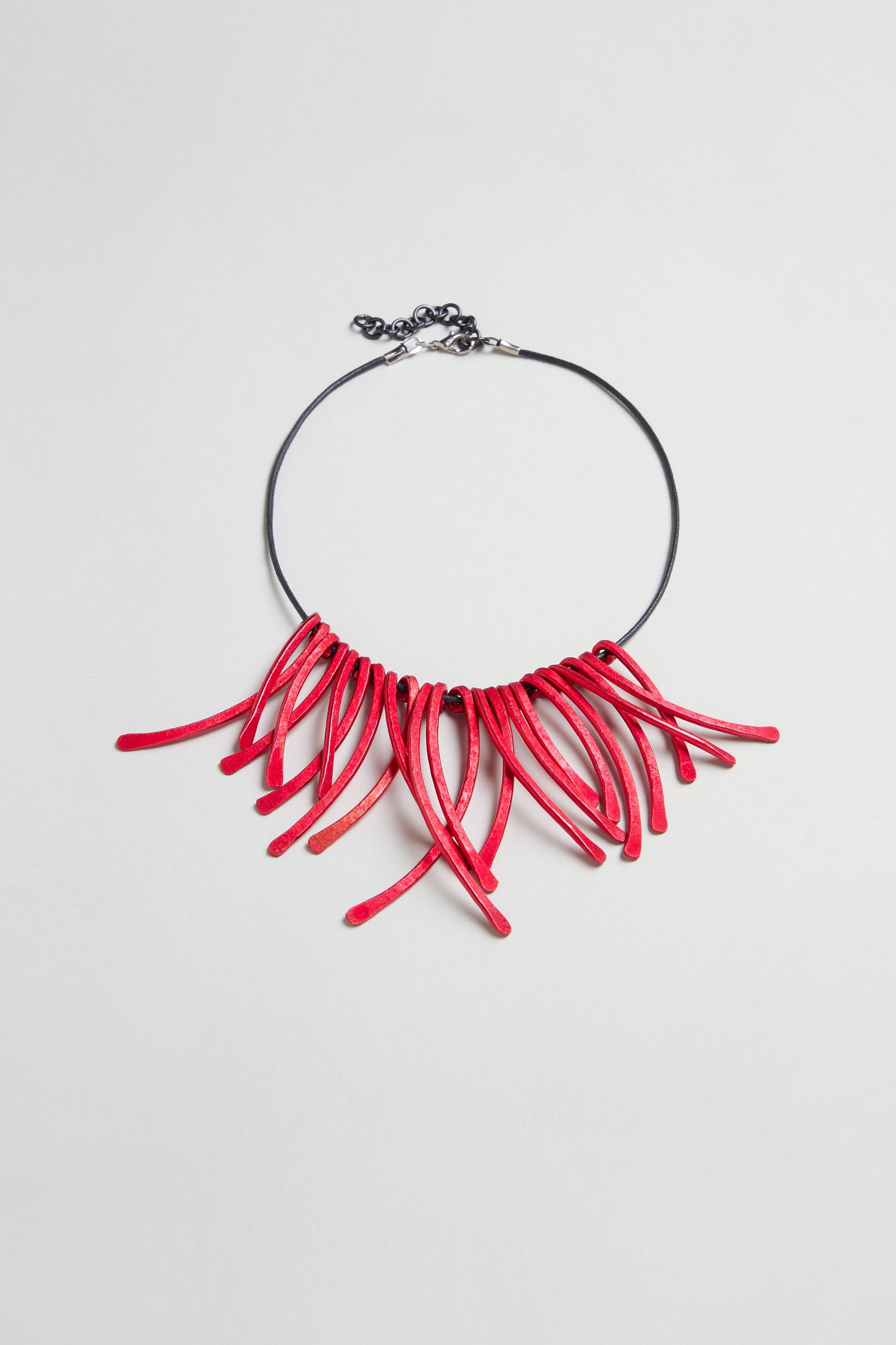 Introducing the Arc Necklace: a black choker featuring multiple curved red anodised aluminium elements fanned out at the front, secured with an adjustable metal clasp closure. This contemporary jewellery piece is crafted as wearable art, making a bold and striking statement.