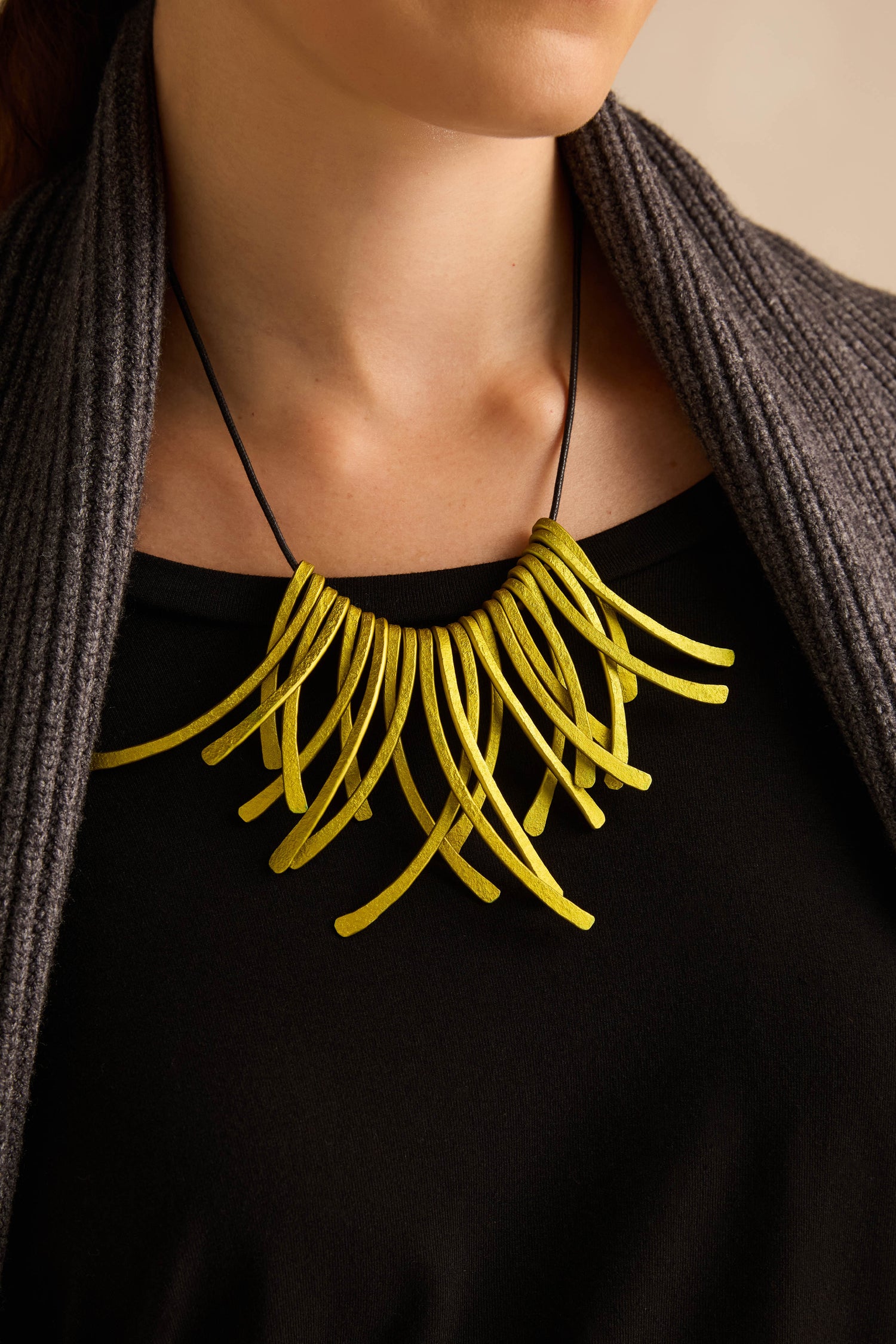 A person wearing a black top and gray cardigan, showcasing the Arc Necklace—an eye-catching piece of contemporary jewellery crafted from bold yellow anodised aluminium with multiple curved, tube-like elements that double as wearable art.