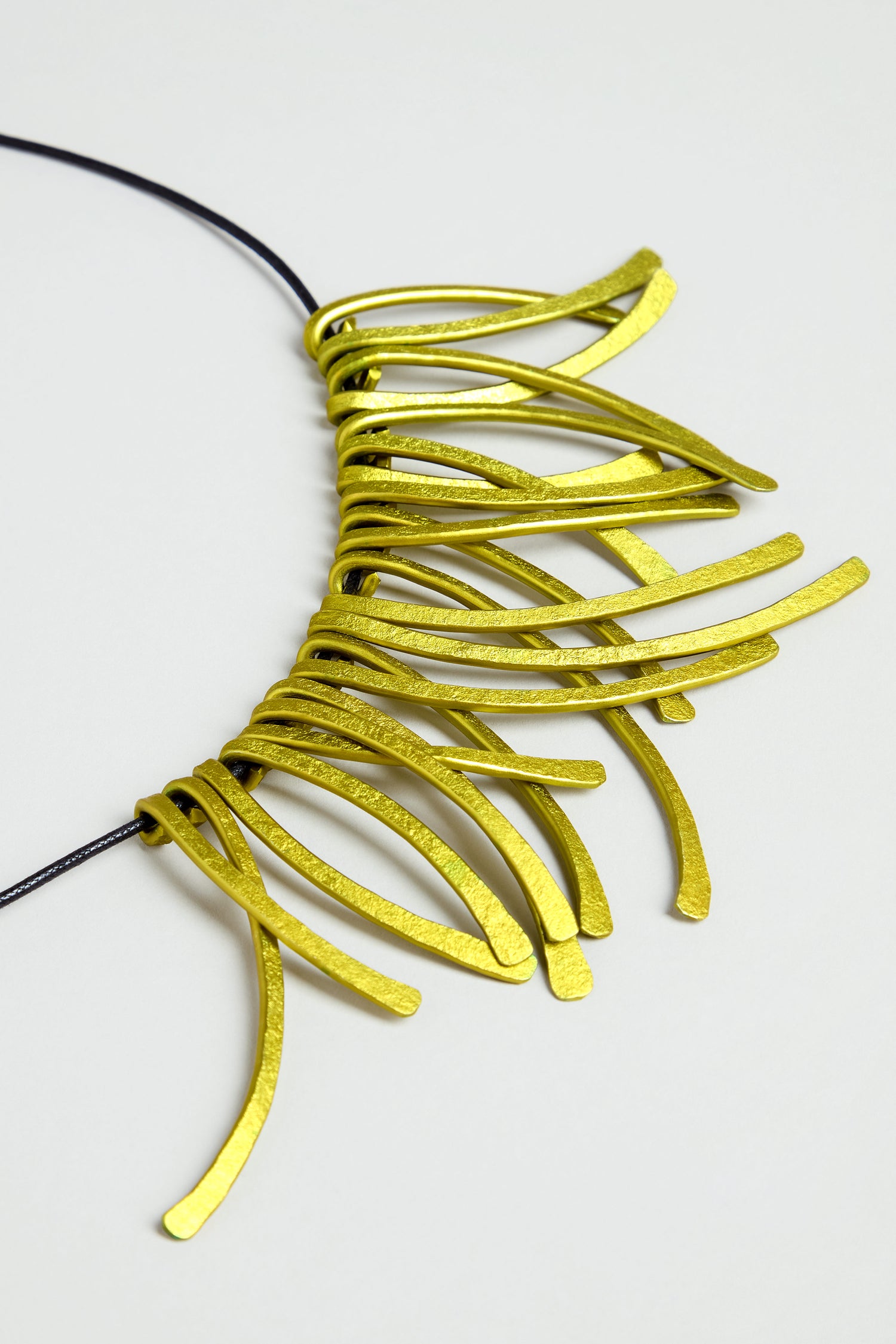 Introducing the Arc Necklace: a contemporary piece crafted from multiple curved, flat yellow anodised aluminium elements, artfully arranged in a striking fringe pattern along a black cord. Displayed on a light background, this bold yet elegant necklace embodies wearable art through its modern design.