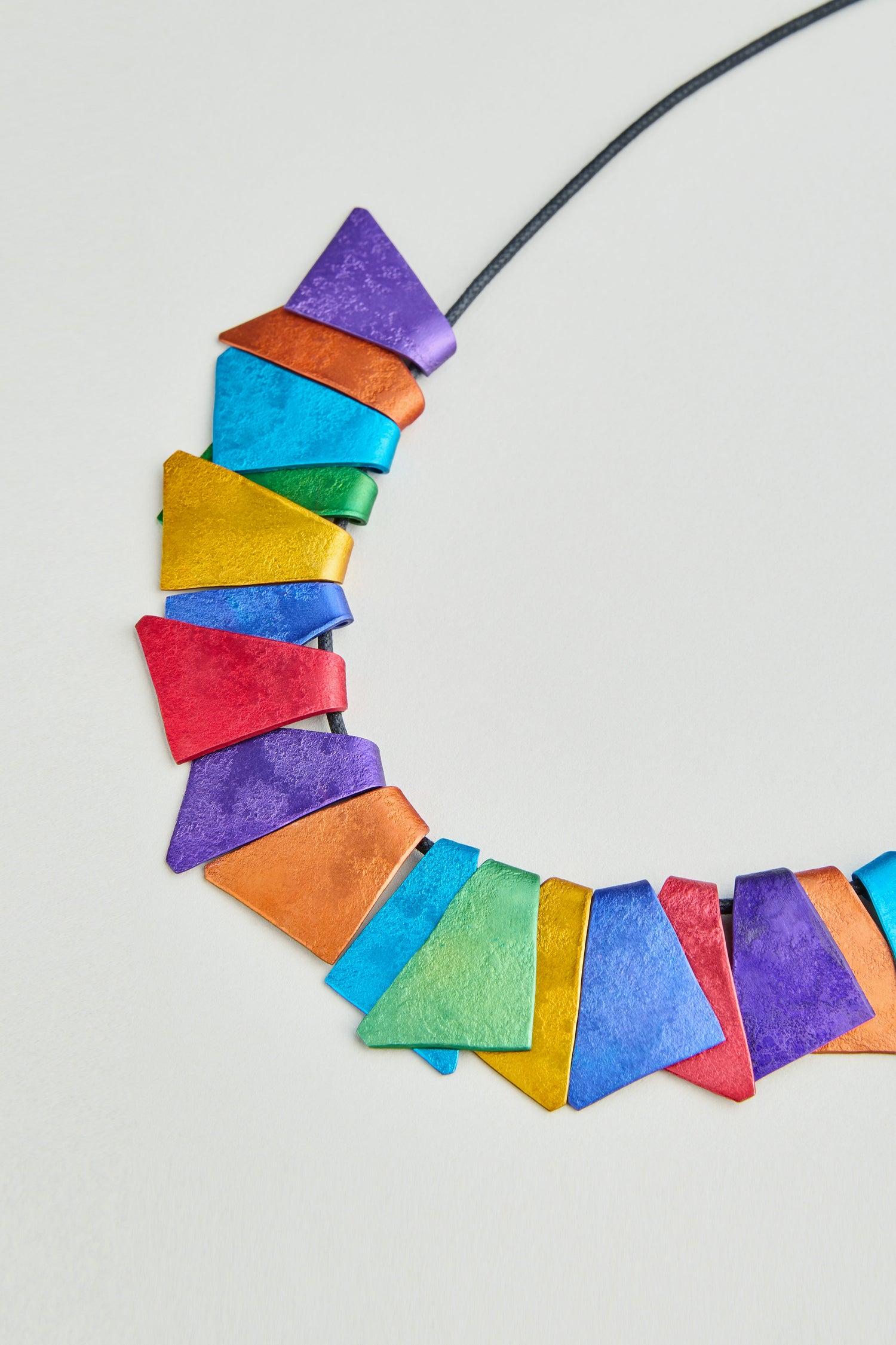 The Mini Segments Necklace features colorful anodised aluminium triangles in red, orange, yellow, green, blue, and purple on a black cord—an eye-catching piece of geometric jewelry displayed on a light background.