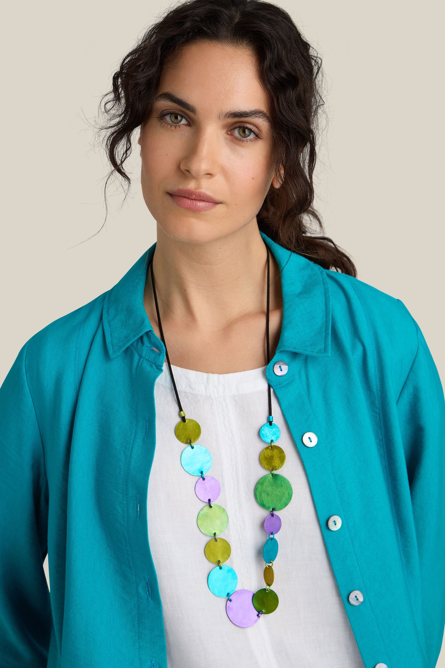 A person with wavy dark hair wears a teal shirt over a white top, accessorized with an Oana Millet handcrafted Colour Pop Circles Necklace featuring circular, multicolored pendants.