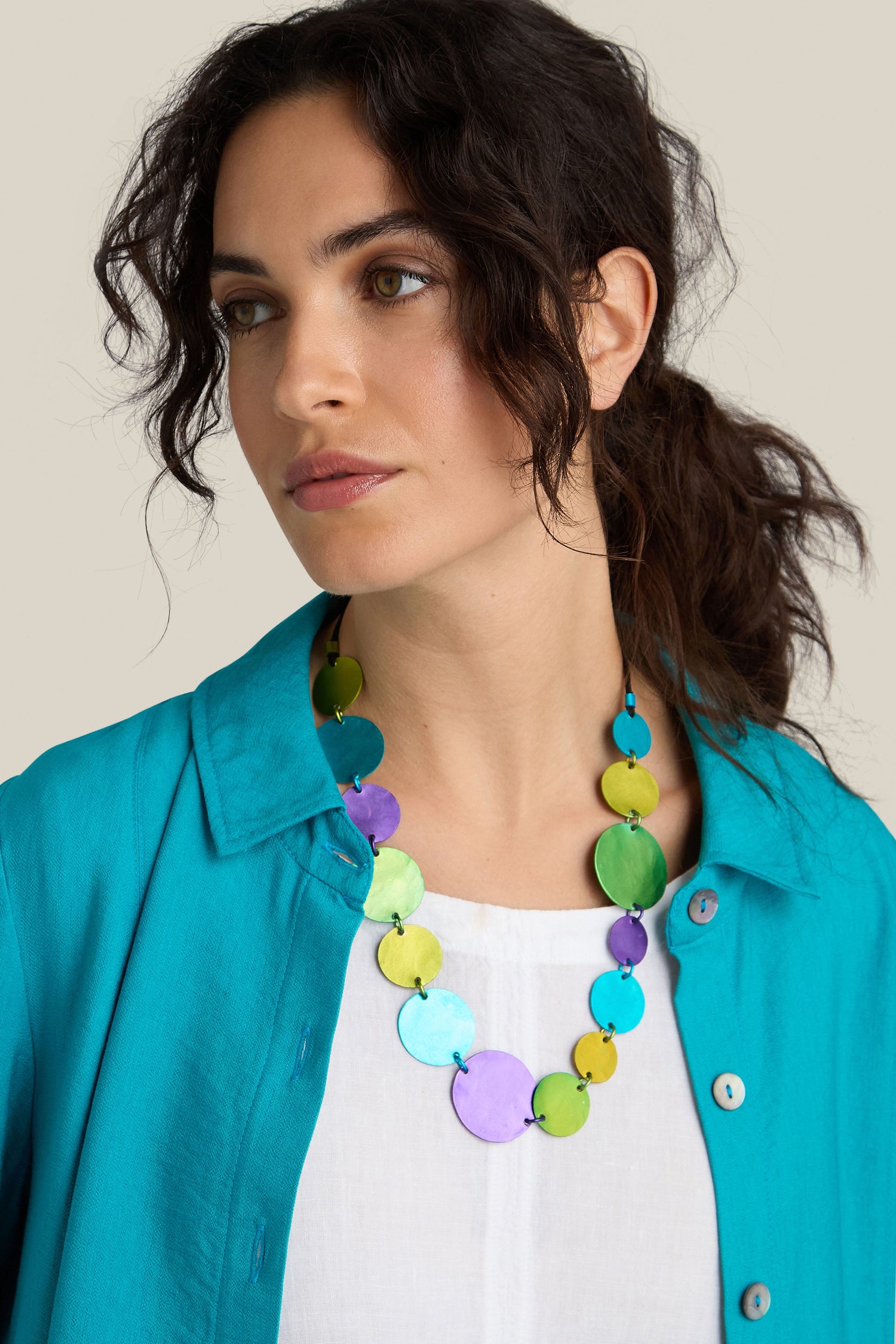 A woman with wavy dark hair, wearing a blue shirt over a white top and an Oana Millet handcrafted Colour Pop Circles Necklace embellished with colorful round beads, looks off to the side.
