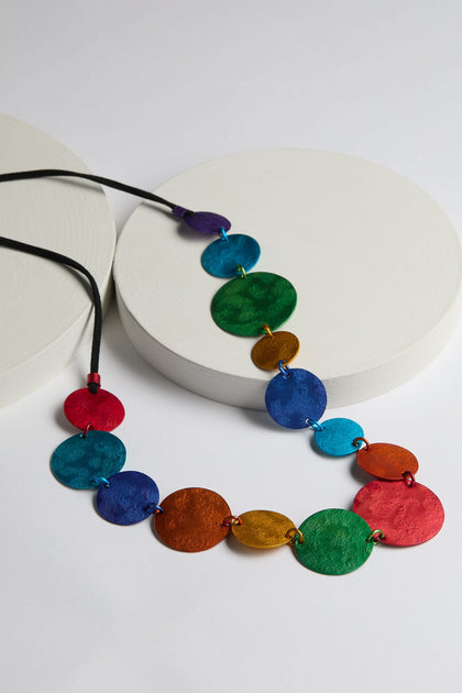 The Colour Pop Circles Necklace features colorful circular pendant with multicolored aluminum discs arranged in a gradient pattern, elegantly positioned on white circular platforms. This exquisite handcrafted necklace is part of the Oana Millet collection.