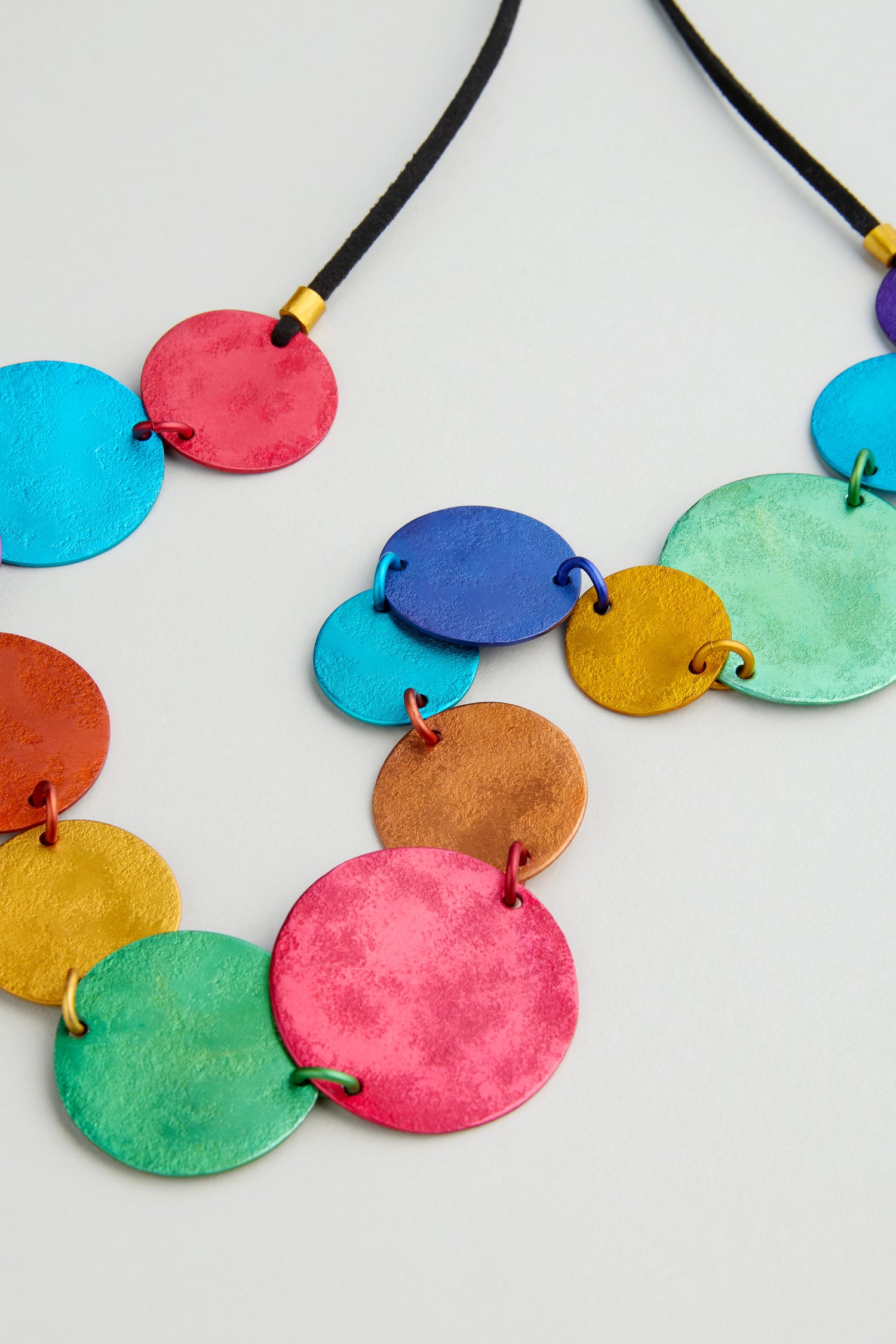 The Colourful Aluminium Discs Necklace features large, flat circular pendants on a black cord. Adjustable in length, it adds a bold, vibrant touch to any outfit and stands out beautifully against a light background.