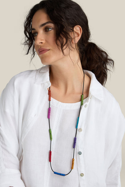 A person with dark wavy hair wearing a white blouse and the Rainbow Spirals Necklace looks to the side against a plain beige background.