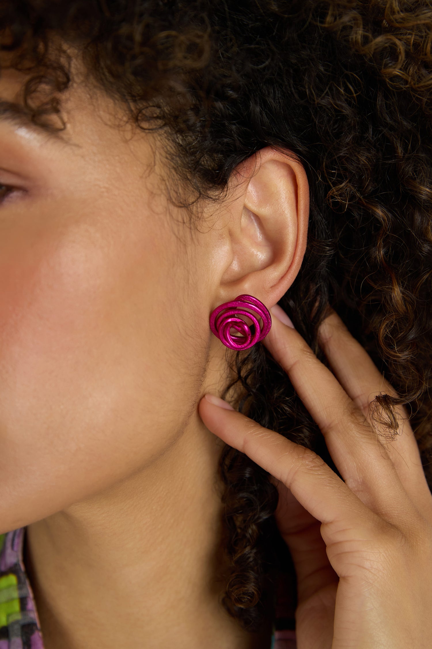 Close-up of a woman with curly hair wearing bright pink Mini Spiral Earrings, crafted from anodised aluminium for a modern look. Her hand gently touches her ear, highlighting the sculptural design.