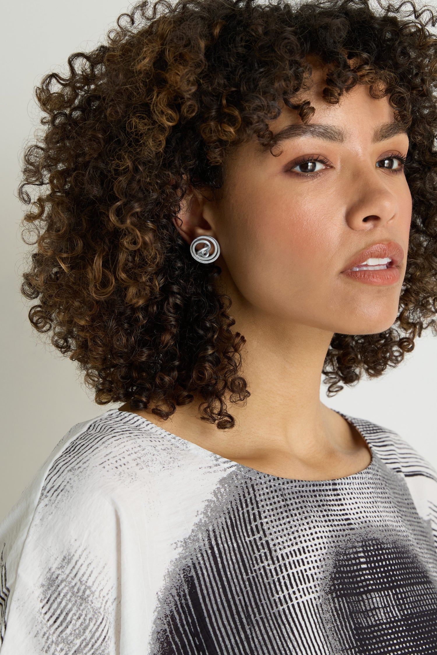 A woman with curly hair in a patterned top and sculptural earrings looks to the side with a neutral expression, highlighting bold Mini Spiral Earrings.