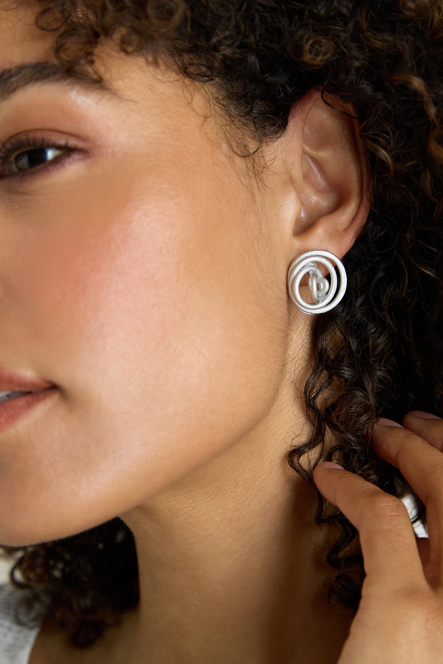 Close-up of a woman wearing Mini Spiral Earrings—sculptural earrings made from anodised aluminium—as she gently touches her curly hair.