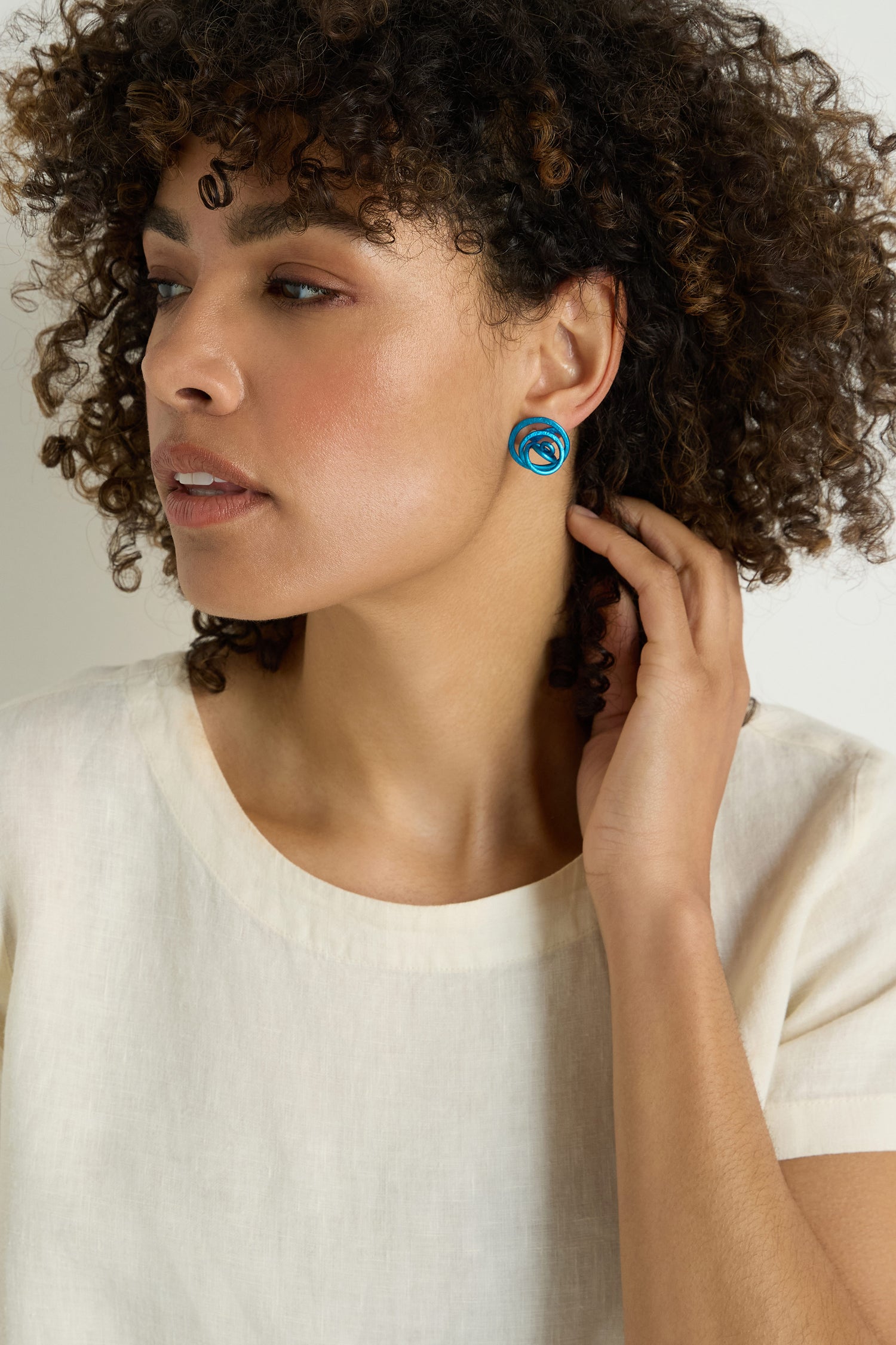 A woman with curly hair, wearing a cream top and blue Mini Spiral Earrings, looks to the left while touching her ear.