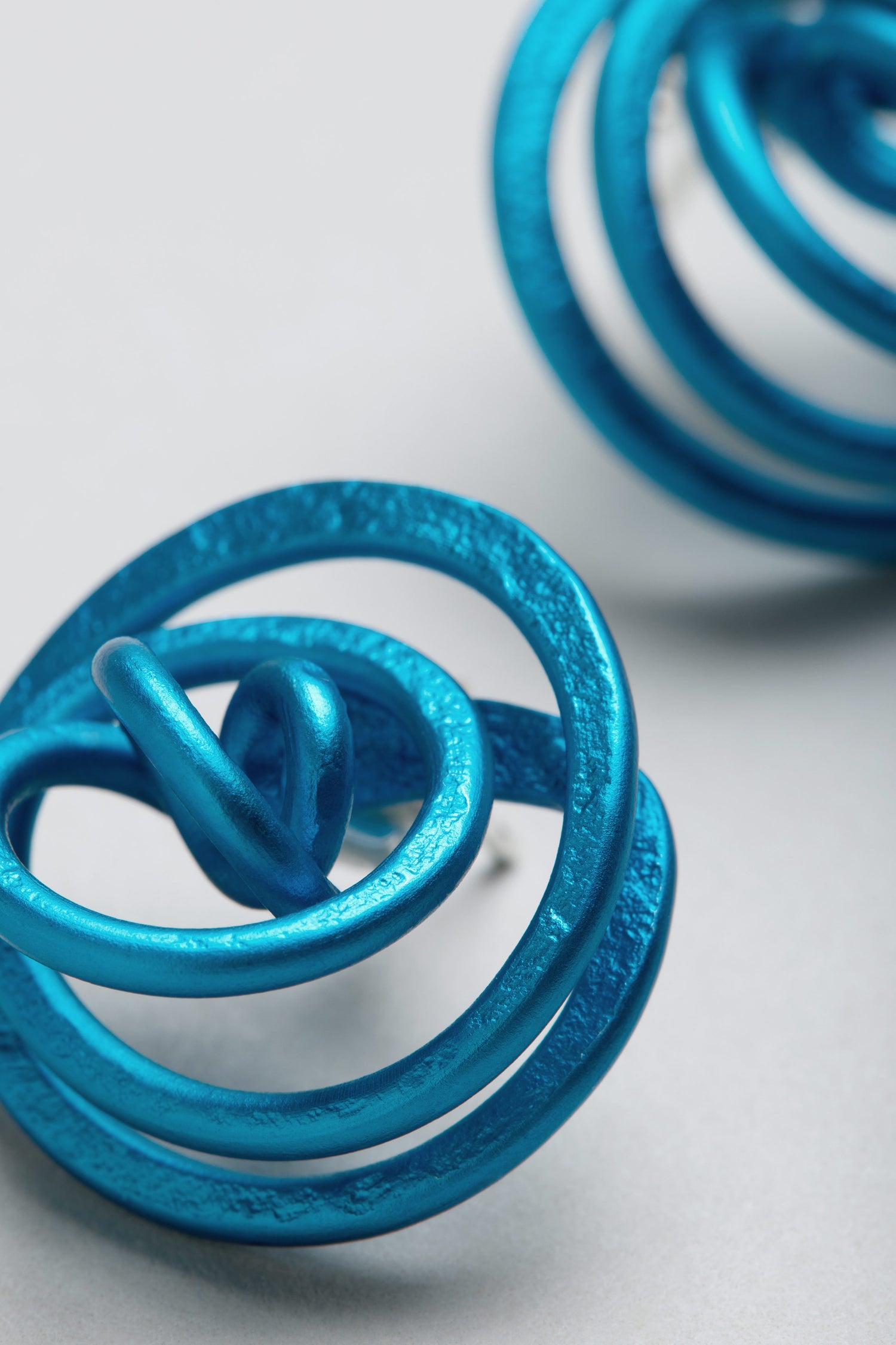 Close-up of two metallic blue Mini Spiral Earrings made from anodised aluminium, displayed on a light gray background.