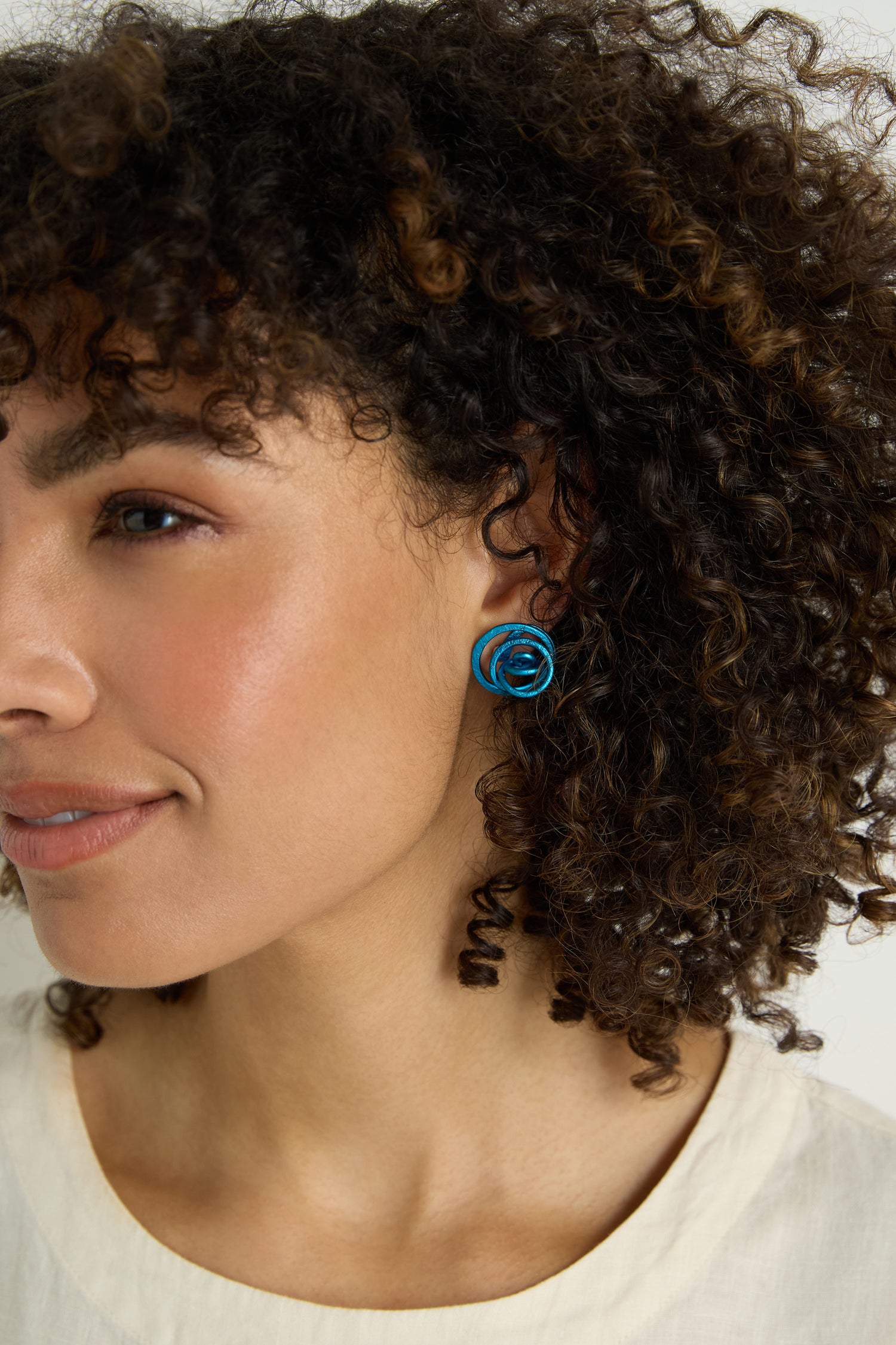 A person with curly hair wears a cream-colored top and shiny blue Mini Spiral Earrings, shown from the cheekbone up against a neutral background.