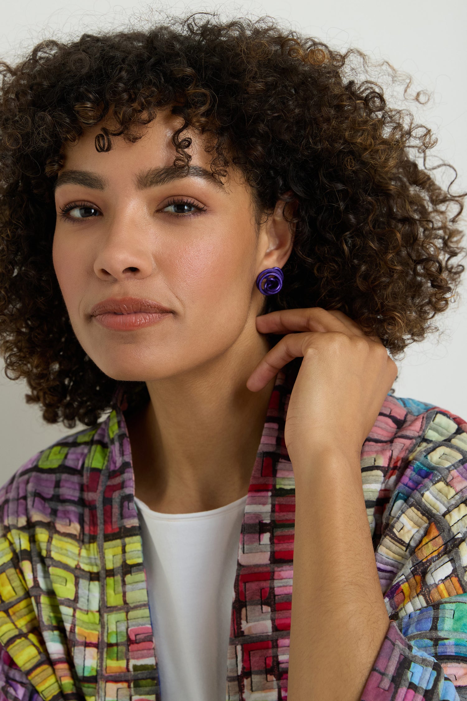 A woman with curly hair, in a colorful patterned jacket and white top, wears the Mini Spiral Earrings and gazes at the camera with a neutral expression.