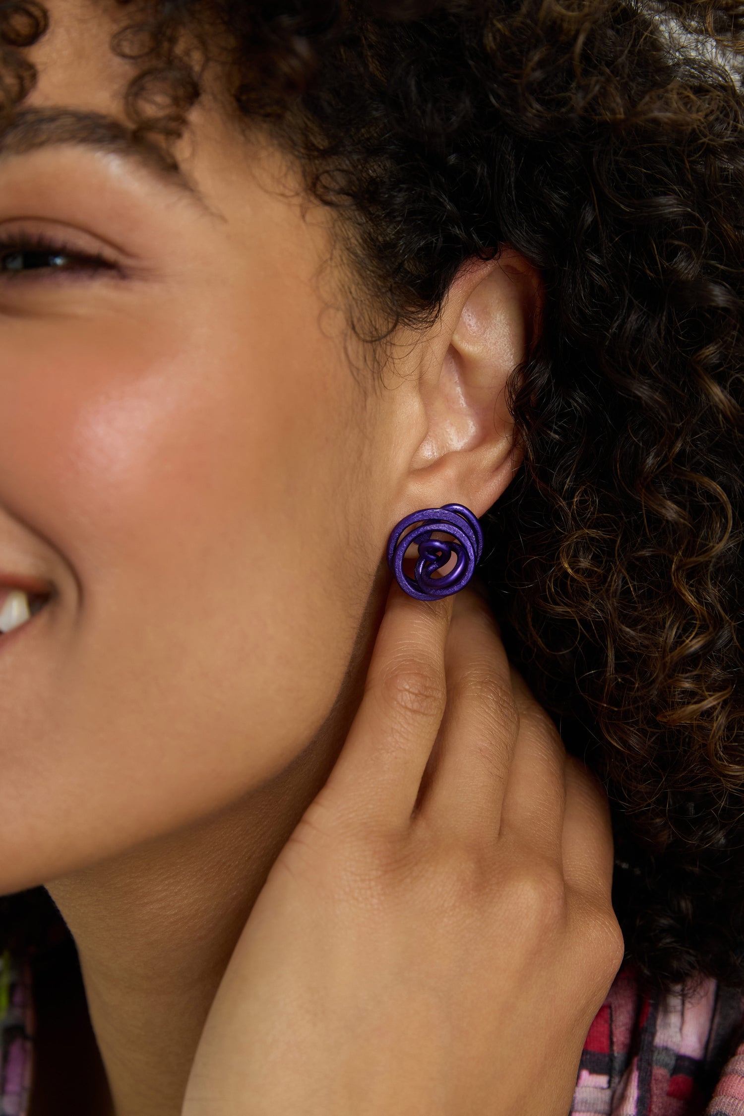 A person with curly hair smiles and gently touches their Mini Spiral Earrings, which feature a sculptural purple rose design.