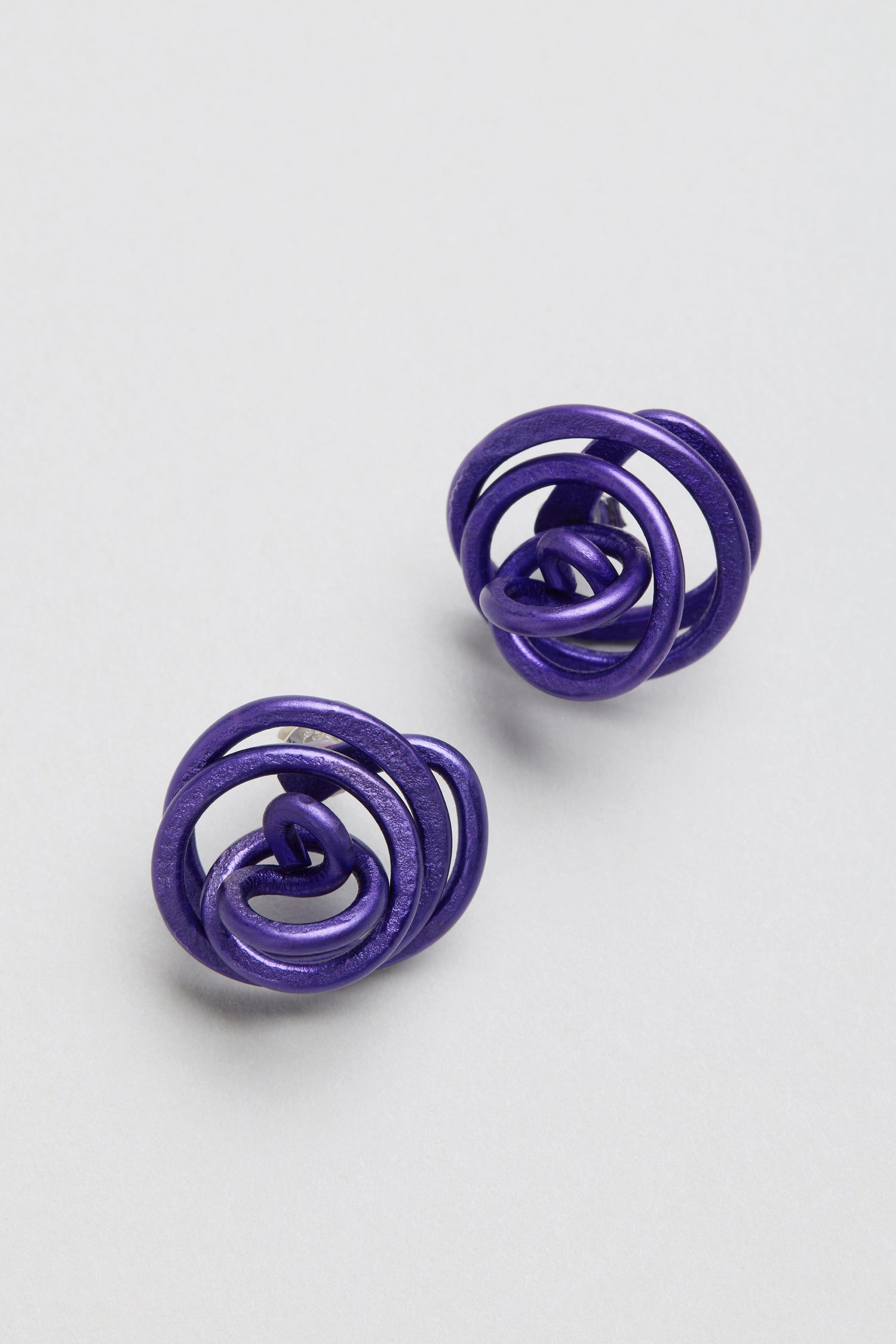 A pair of Oana Millet Mini Spiral Earrings are shown on a light gray background, highlighting their round, purple, abstract and sculptural design.