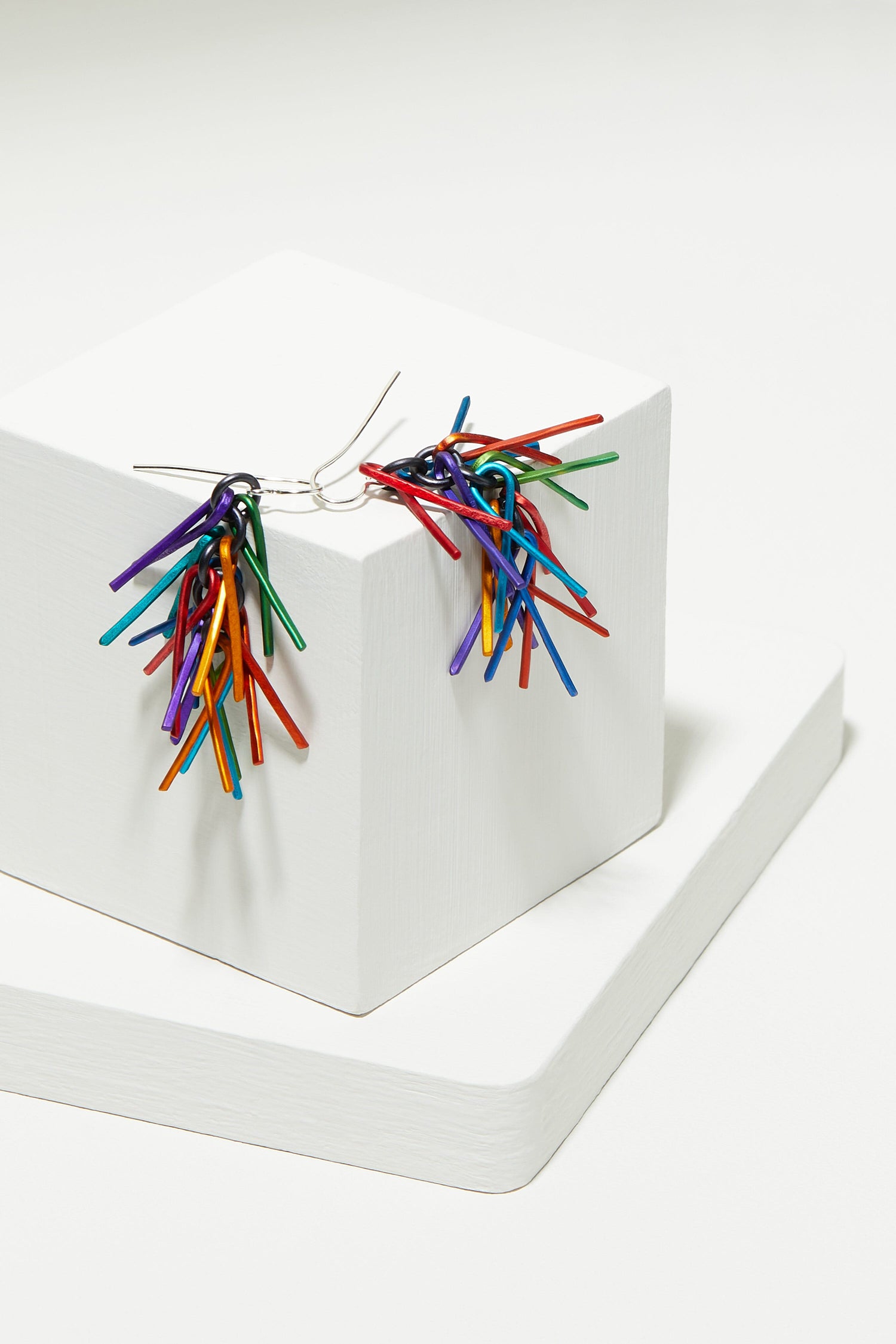 A pair of Fringe Earrings with multi colored twigs on top of a white cube.