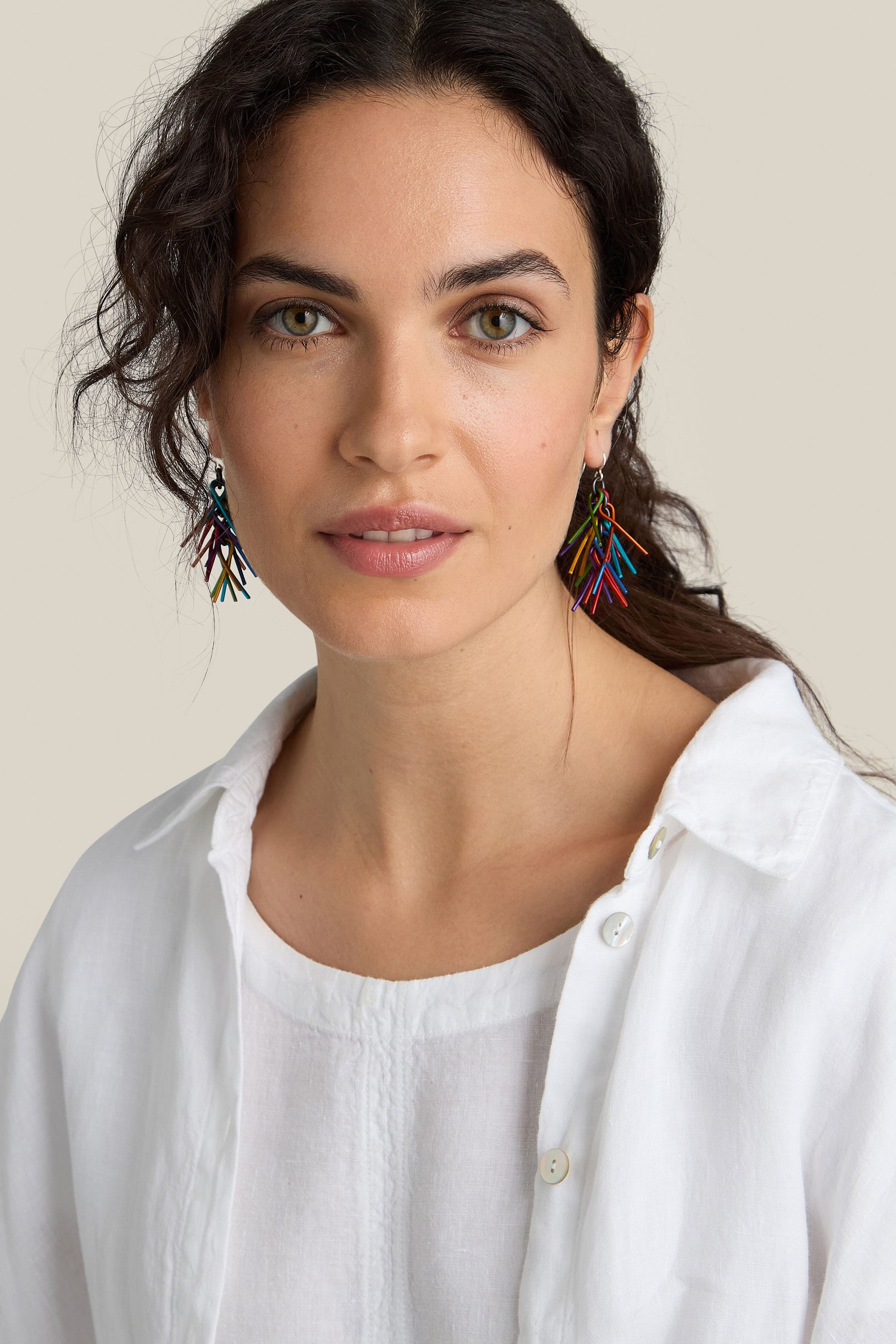 A woman with long dark curly hair is wearing brightly colored Fringe Earrings and a white button-up blouse. She is looking directly at the camera against a neutral background.