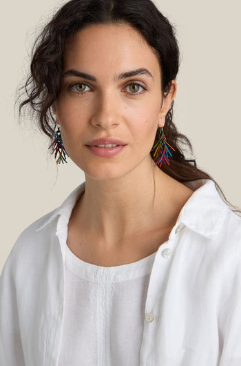 A woman with long dark curly hair is wearing brightly colored Fringe Earrings and a white button-up blouse. She is looking directly at the camera against a neutral background.