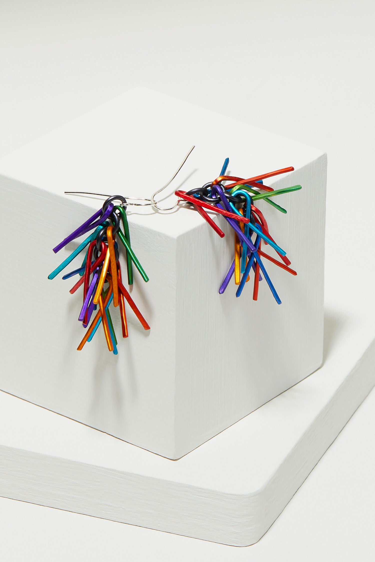 Fringe Earrings, a pair of unique, brightly colored aluminium earrings, displayed on a white geometrical pedestal.