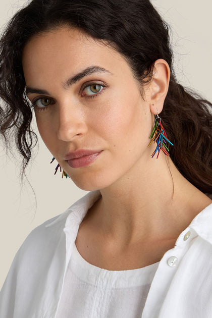 A person with long, wavy dark hair wears unique Fringe Earrings made of brightly coloured aluminium and a white button-up shirt.