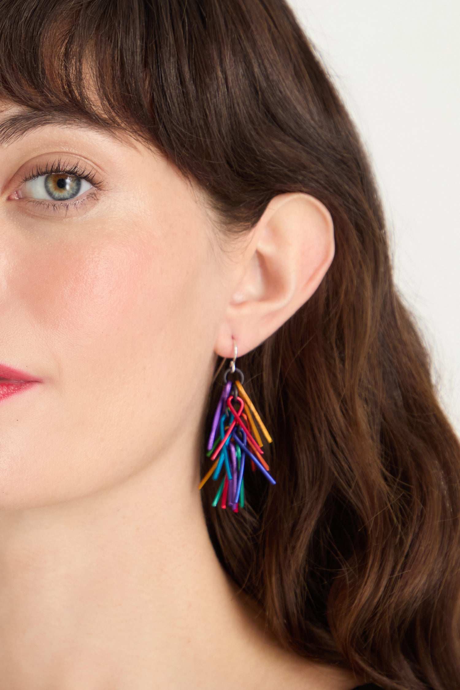 Close-up of a woman with wavy brown hair wearing Fringe Earrings, featuring several thin, curved pieces for a vibrant look. Only the left side of her face is visible.