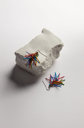 The Fringe Earrings, featuring multicoloured aluminium spears, are displayed—one on a rough white stone and the other on a white surface—highlighting their unique craftsmanship.