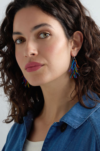 Woman with wavy dark hair wears Fringe Earrings—colourful aluminium jewellery styled as multicoloured spears—with a blue denim jacket, gazing slightly to the side against a plain background.