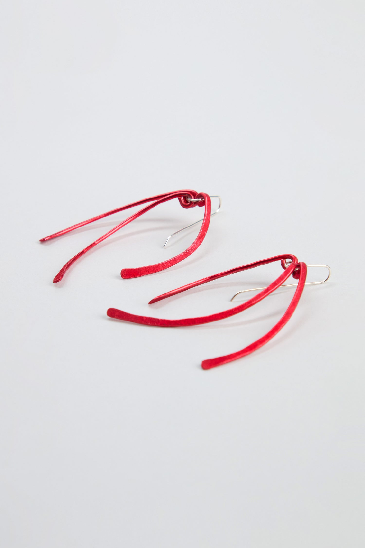Introducing the Arc Earrings: a pair of elongated, curved earrings in vibrant red anodised aluminium with sterling silver hooks, showcased against a simple light background.