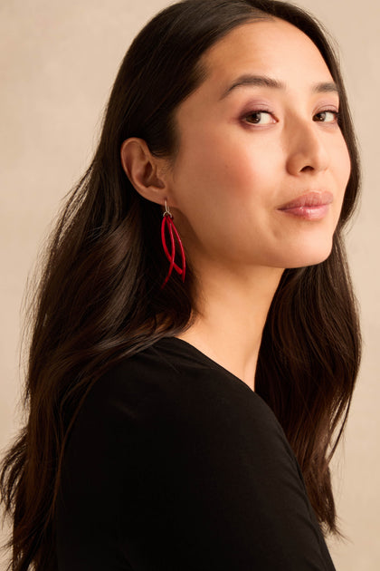 A woman with long dark hair wearing a black top showcases the Arc Earrings—striking red earrings with sterling silver hooks—as she looks at the camera against a plain beige background.