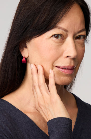 A woman with long dark hair wearing a dark top and bold pink Curved Earrings looks to her left against a neutral background.
