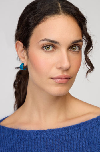 A woman with wavy dark hair, wearing Curved Earrings and a blue knit sweater, looks directly at the camera against a plain background.
