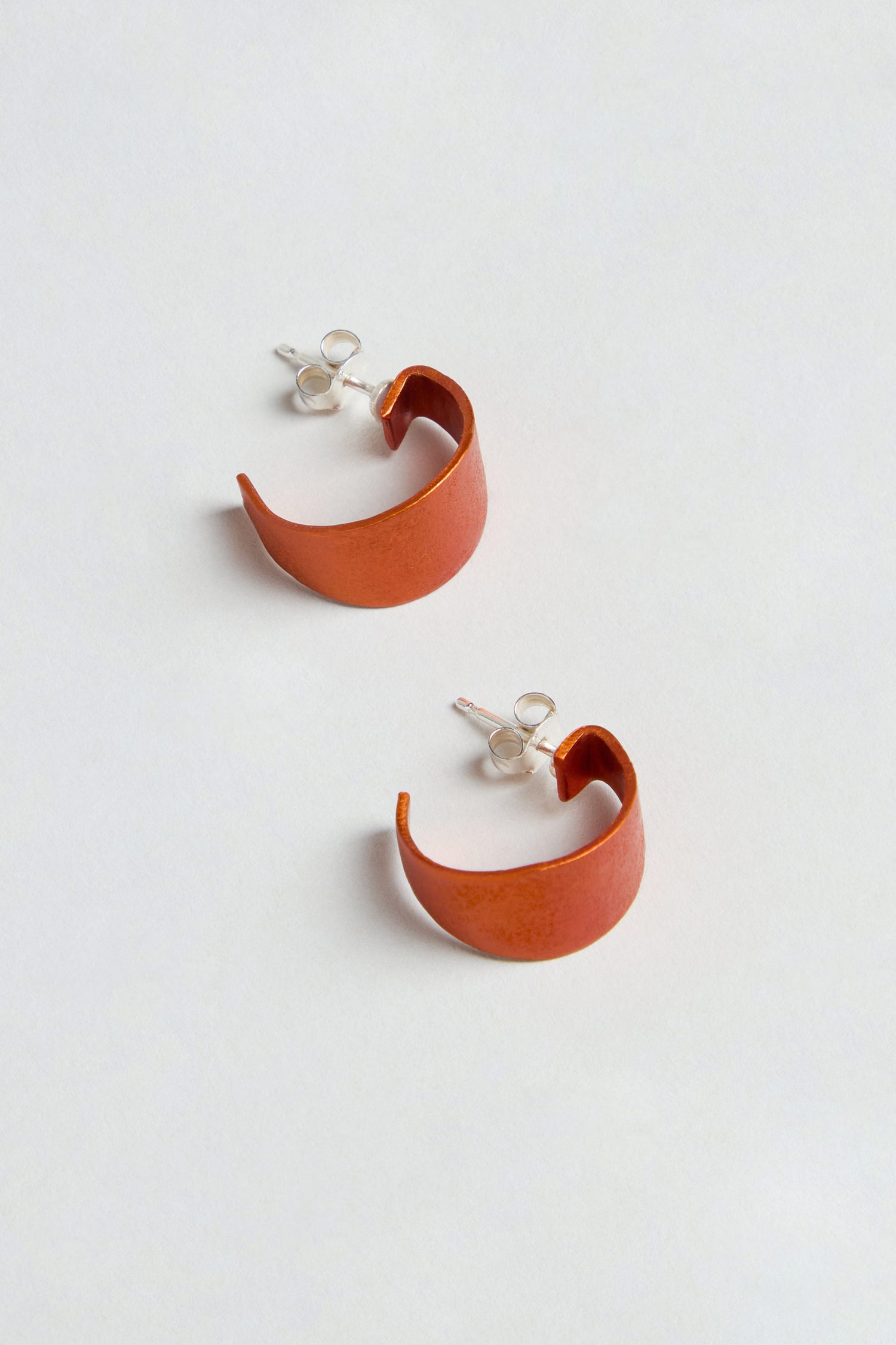 Curve Earrings: A pair of orange, crescent-shaped earrings made from anodised aluminium with sterling silver posts, shown on a light surface.