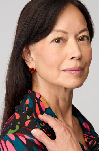 A woman with straight dark hair wears a patterned shirt and Ribbon Earrings made of red anodised aluminium, looking at the camera with a neutral expression and her hand resting on her shoulder.