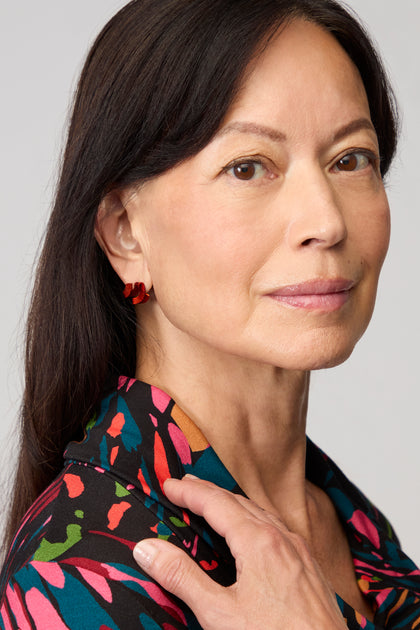 A woman with straight dark hair wears a patterned shirt and Ribbon Earrings made of red anodised aluminium, looking at the camera with a neutral expression and her hand resting on her shoulder.