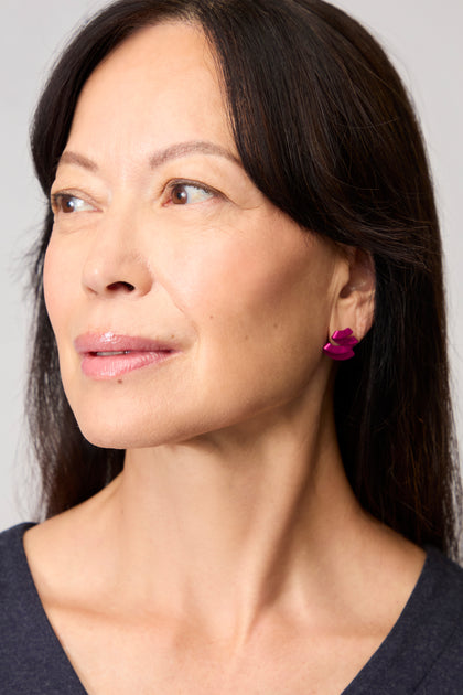 A woman with long dark hair looks to the side, wearing a dark top and pink Ribbon Earrings featuring a sculptural, flower-shaped anodised aluminium design.