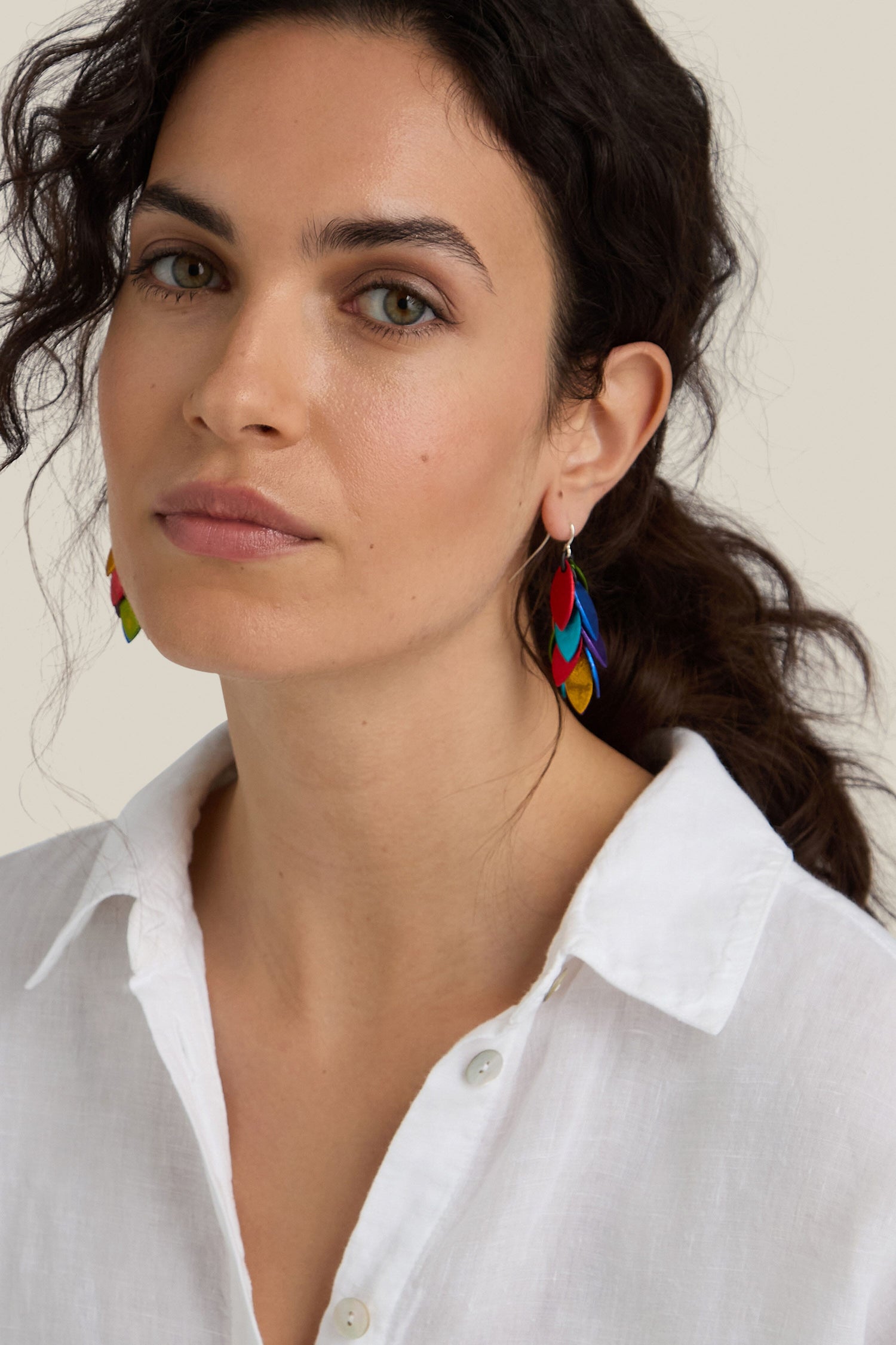 A woman with wavy dark hair wears a white button-up shirt and unique, colorful Scattered Leaves Earrings from a Spanish design studio. She looks into the camera with a neutral expression.