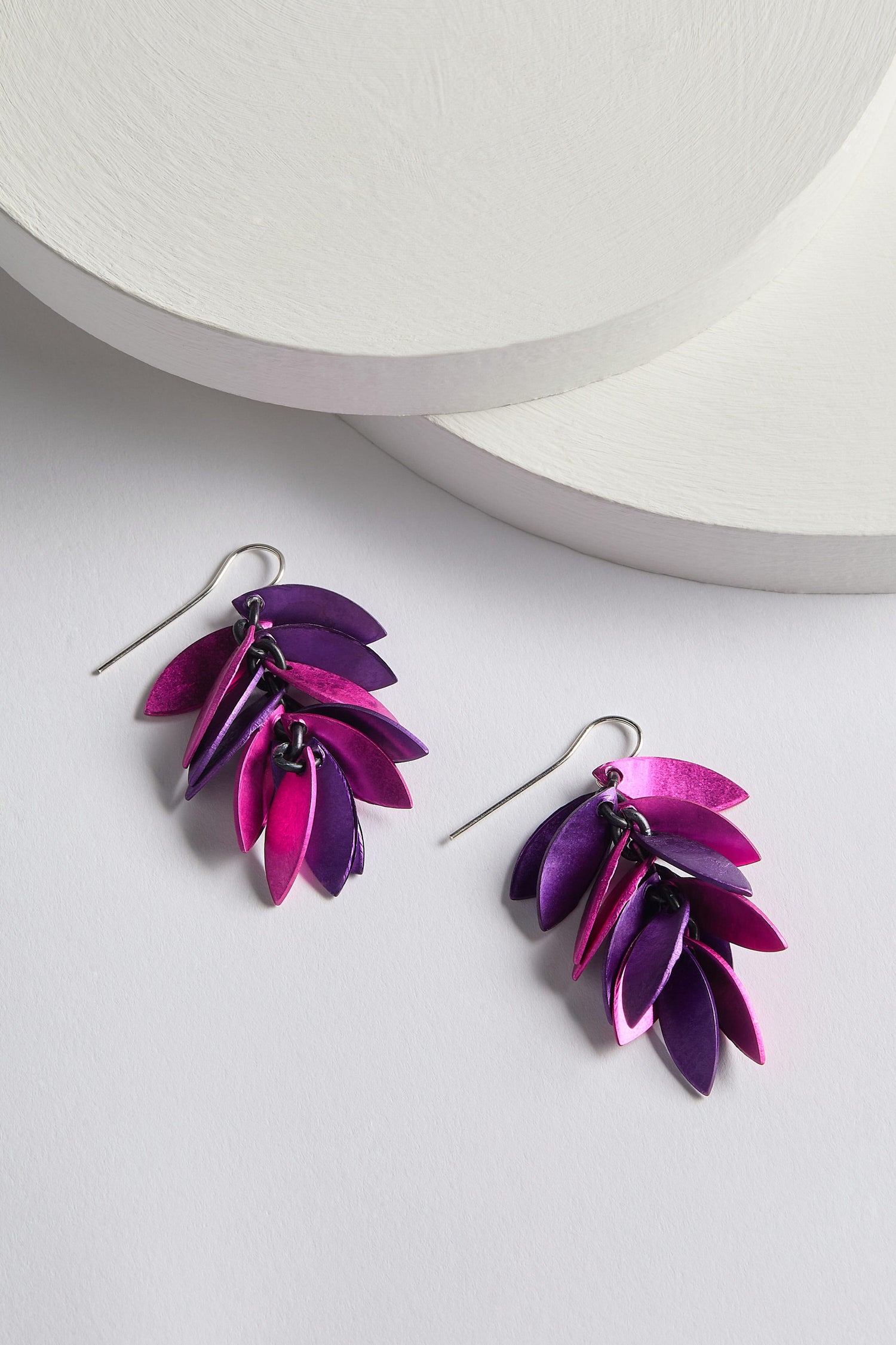 The Scattered Leaves Earrings, featuring layered purple and pink leaf-like shapes arranged in a cascading design, are crafted by a Spanish design studio using sophisticated aluminium jewellery techniques. They are elegantly displayed on a light surface with circular objects in the background.