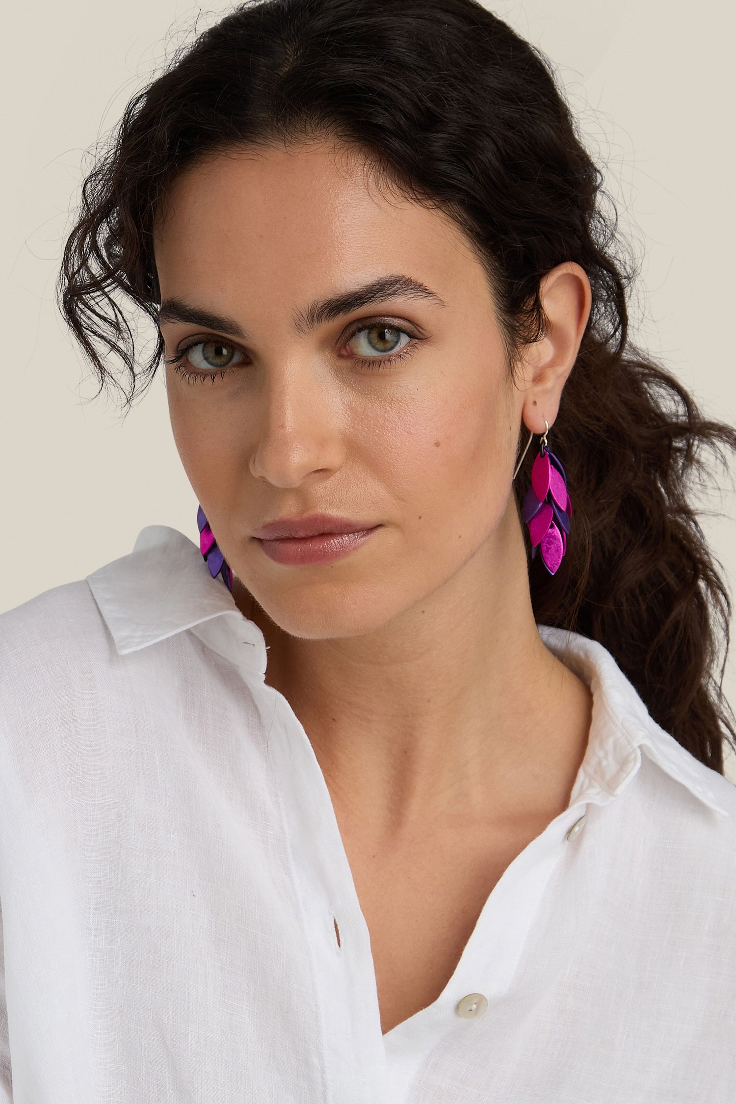 A person with long dark hair wearing the unique Scattered Leaves Earrings, which are bright purple and pink feather-shaped pieces made from aluminium jewellery, paired perfectly with a white button-up shirt.