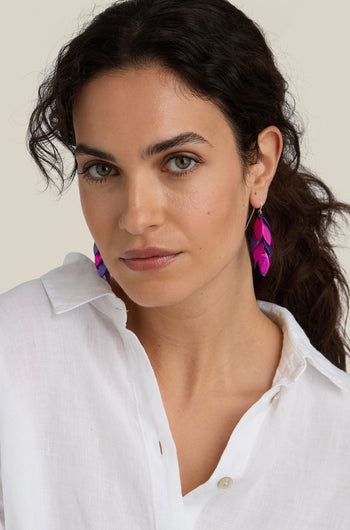 A person with long dark hair wearing the unique Scattered Leaves Earrings, which are bright purple and pink feather-shaped pieces made from aluminium jewellery, paired perfectly with a white button-up shirt.