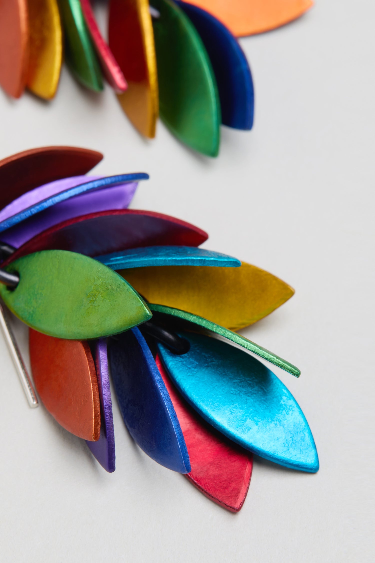 Close-up of overlapping, leaf-shaped pieces in metallic green, blue, red, orange, and purple on a light background, inspired by the Scattered Leaves Earrings.