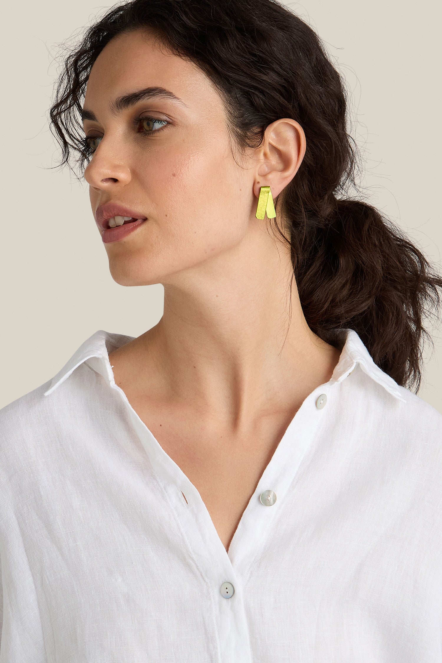 A woman with dark hair in a ponytail is wearing a white collared shirt and vibrant Origami Earrings from a Spanish design studio. She looks to the side against a plain background.