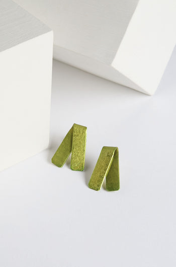 A pair of Origami Earrings in vibrant green metallic, featuring a folded rectangular design, placed on a white surface near two white geometric shapes, crafted by a renowned Spanish design studio.