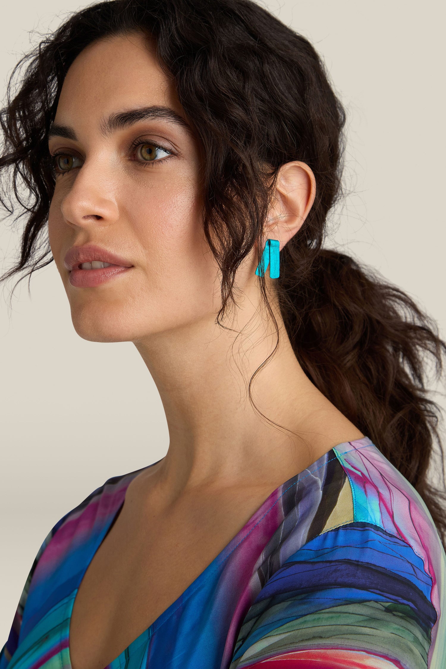 A woman with dark curly hair in a ponytail wears vibrant turquoise Origami Earrings and a multicolored top, looking slightly to the side against a plain background.