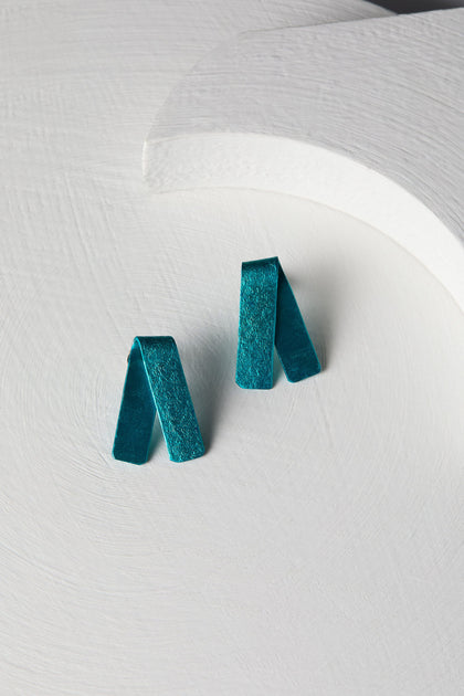 A pair of Origami Earrings in vibrant teal, crafted from folded fabric into rectangular shapes, is placed on a textured white circular surface, showcasing the exquisite craftsmanship of a Spanish design studio.